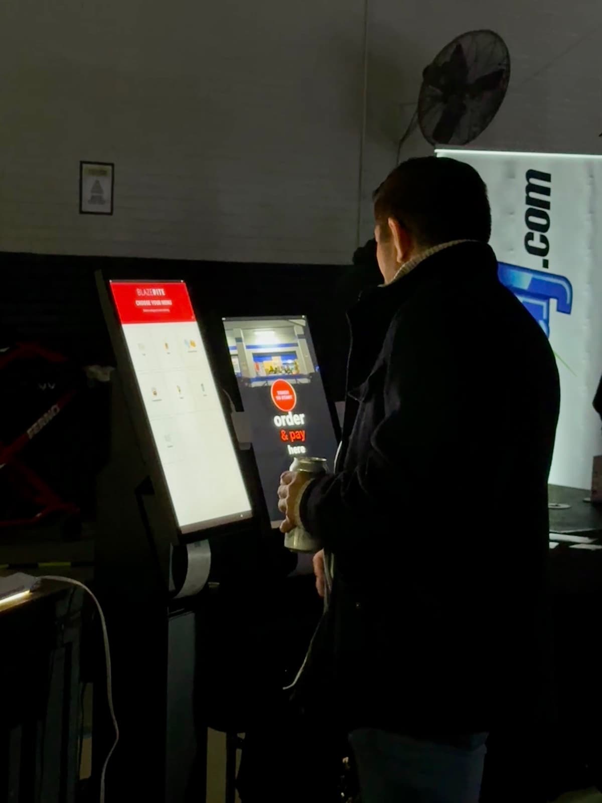 A customer at a BlazeBite self-checkout kiosk, seen from behind, with the order and pay screen lit