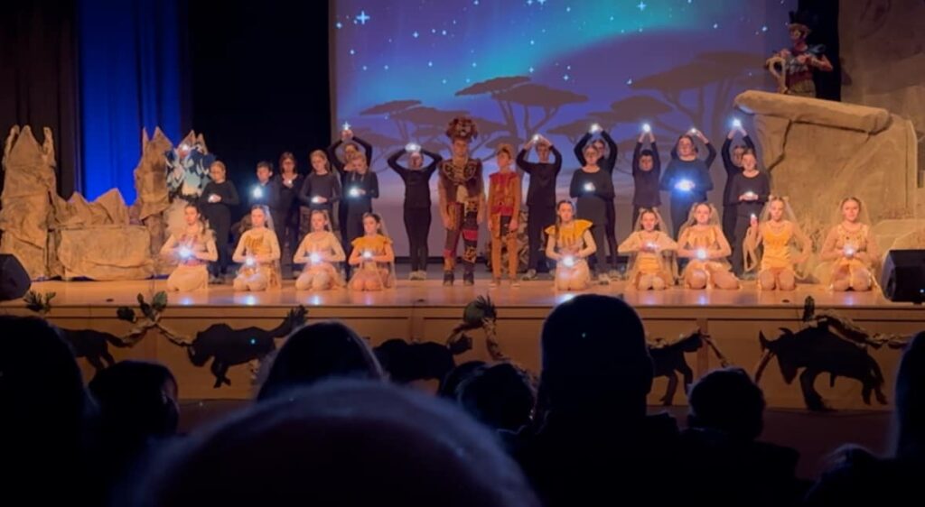 Lion King Jr. cast performing under a night-sky backdrop, each holding a small light onstage near the hand-built Pride Rock