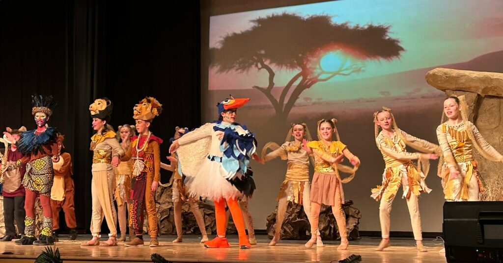 Lion King Jr. cast on stage in custom, hand-crafted costumes and detailed headdresses—some taking over 20 hours to create—built by dedicated parent volunteers.