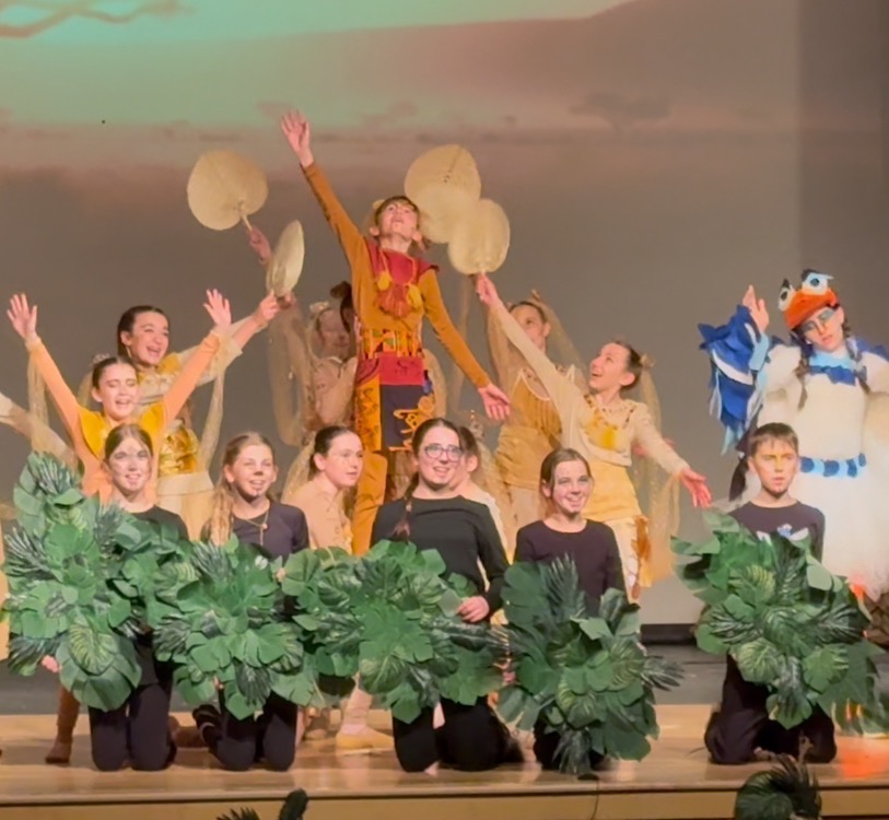 Lion King Jr. cast performing onstage, children raising arms in celebration with custom fans and leaf props, after five months of dedicated rehearsals and parent-crafted costumes.